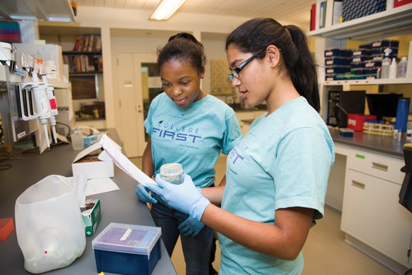 PHOTO: Two high school girls wearing blue "College First" tshirts and latex gloves examine samples in the lab.