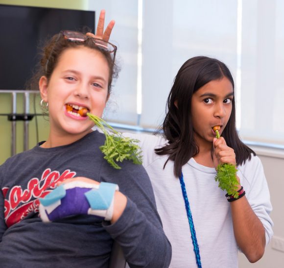 PHOTO: Two girls are eating carrots. One holds two fingers up behind the other's head to give her bunny ears.